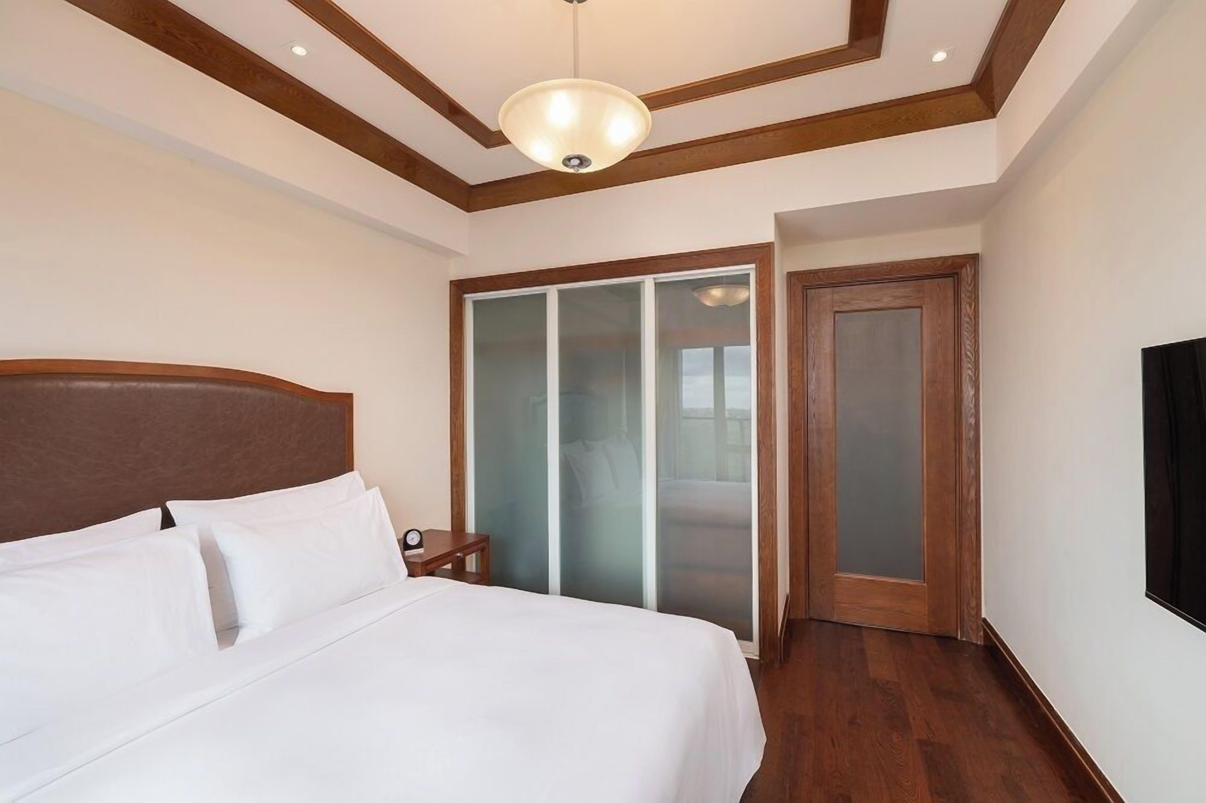 Habitación de hotel con una cama grande, cabecera de madera, mesita de noche, televisor y puerta corrediza de vidrio que da a un balcón.
