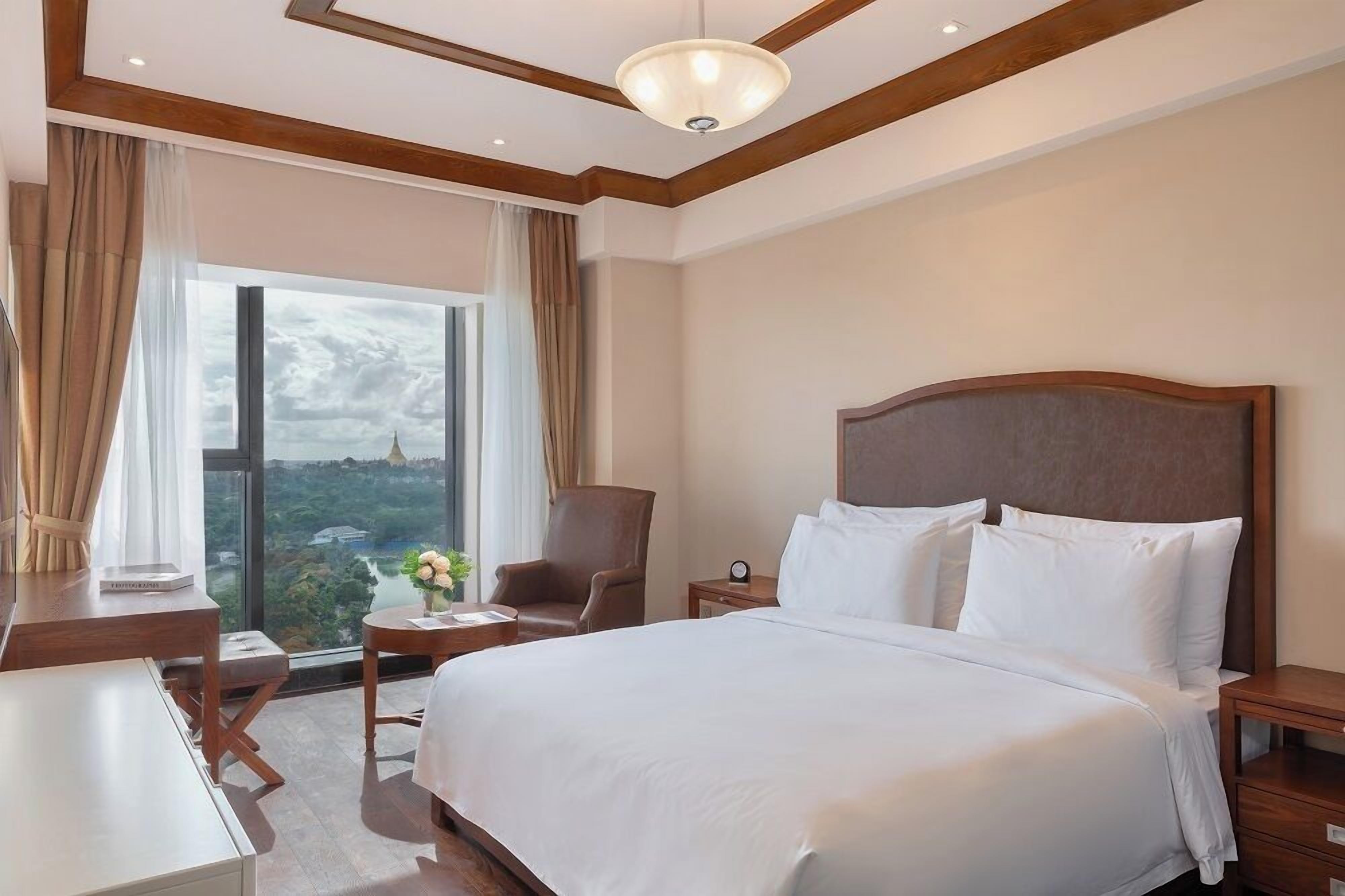 A hotel room with a large bed, a desk, a chair, and a view of a cityscape through large windows.