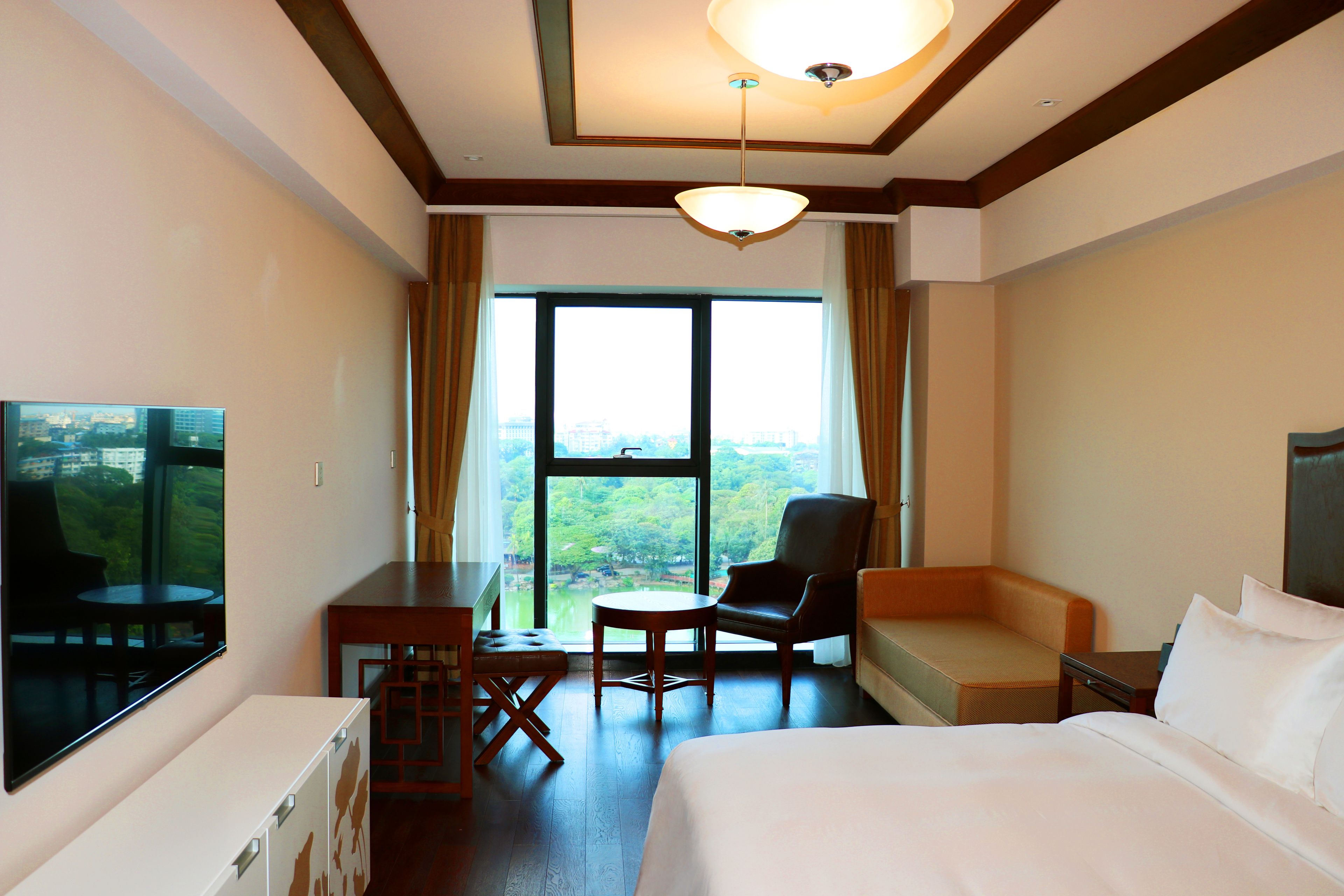 Habitación de hotel con cama, escritorio, silla, televisión y un amplio ventanal con vistas al verdor.