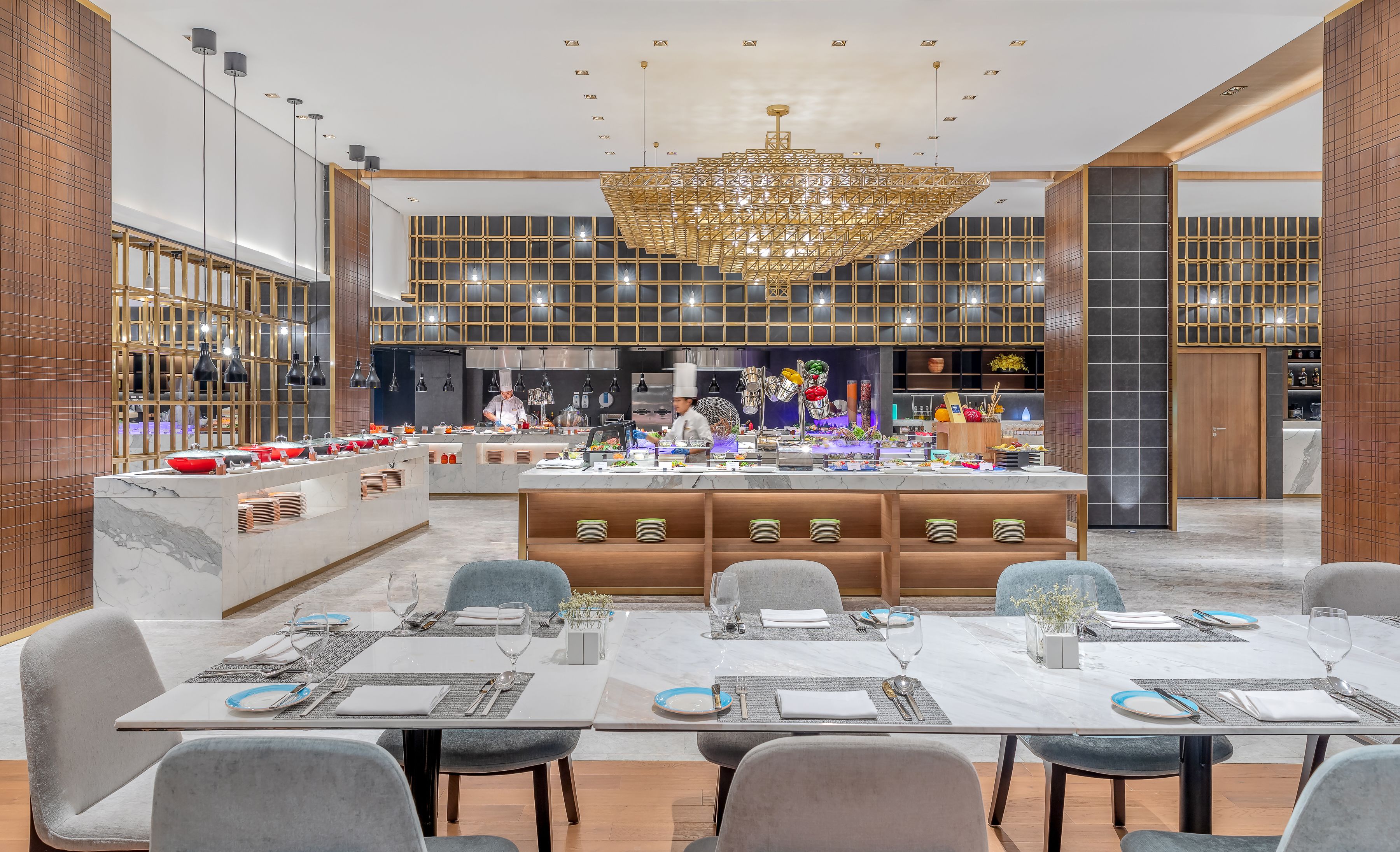 A modern dining area with marble tables, grey upholstered chairs, and a central island with a chef preparing food.