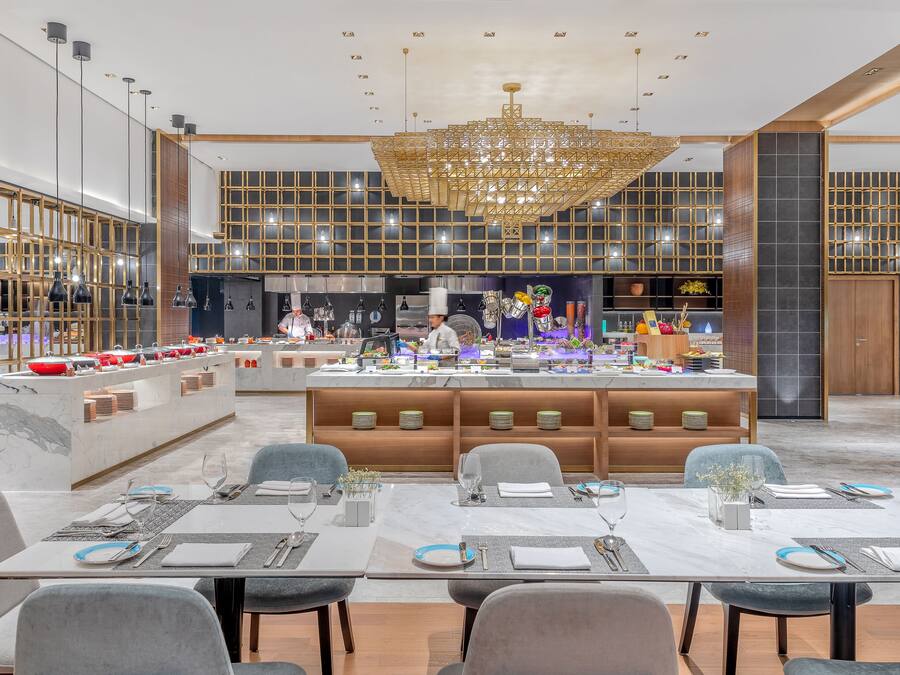 A modern dining area with marble tables, grey upholstered chairs, and a central island with a chef preparing food.