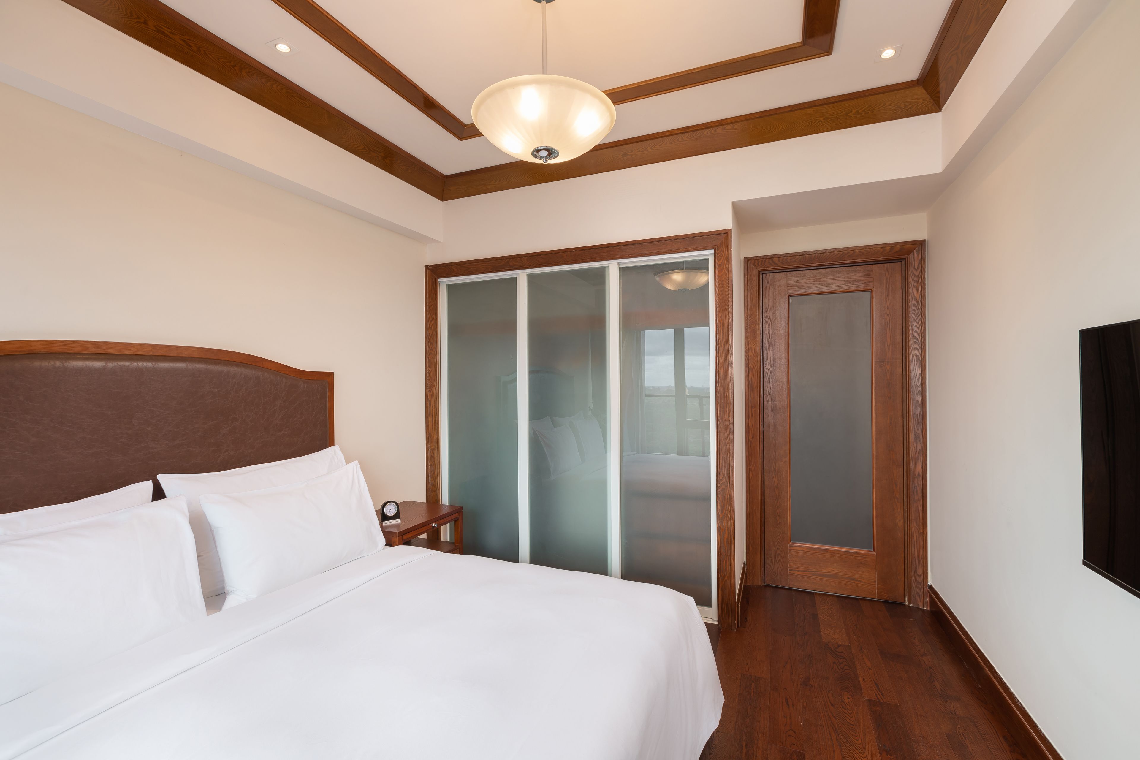 A hotel room with a large bed, wooden headboard, a nightstand, a TV, and a sliding glass door leading to a balcony.