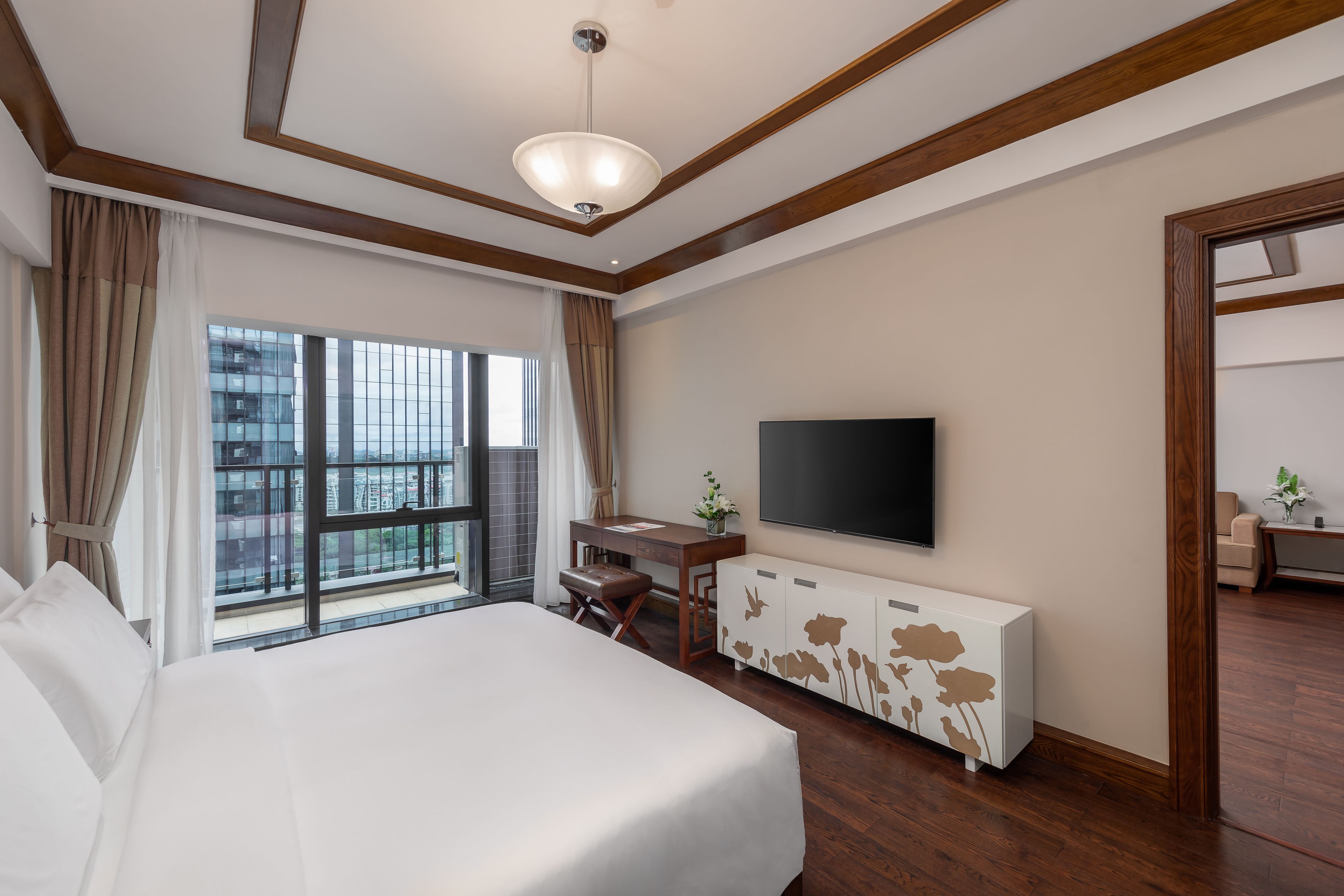 A hotel room with a large bed, a TV mounted on the wall, a desk with a chair, and a view of a cityscape through the window.