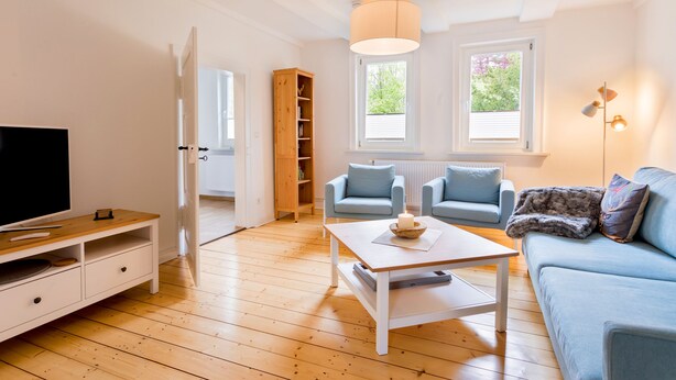 Ein Wohnzimmer mit Fernseher, einer Holz-Unterhaltungsecke, einem Couchtisch, zwei Sofas, einem Bücherregal und einem Fenster mit Jalousien.