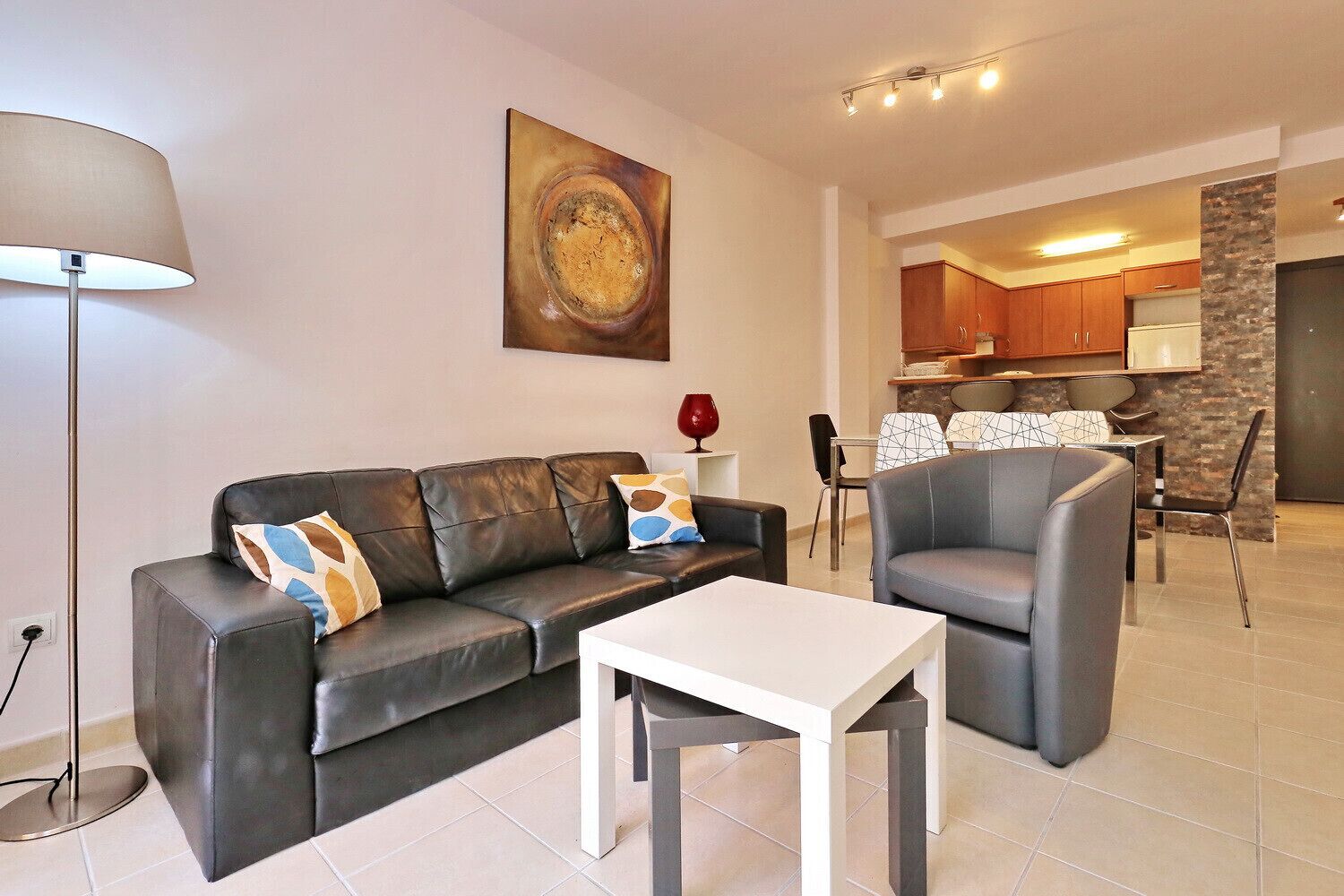 A modern living room with a leather sofa, a white coffee table, and a dining area with a patterned tablecloth.