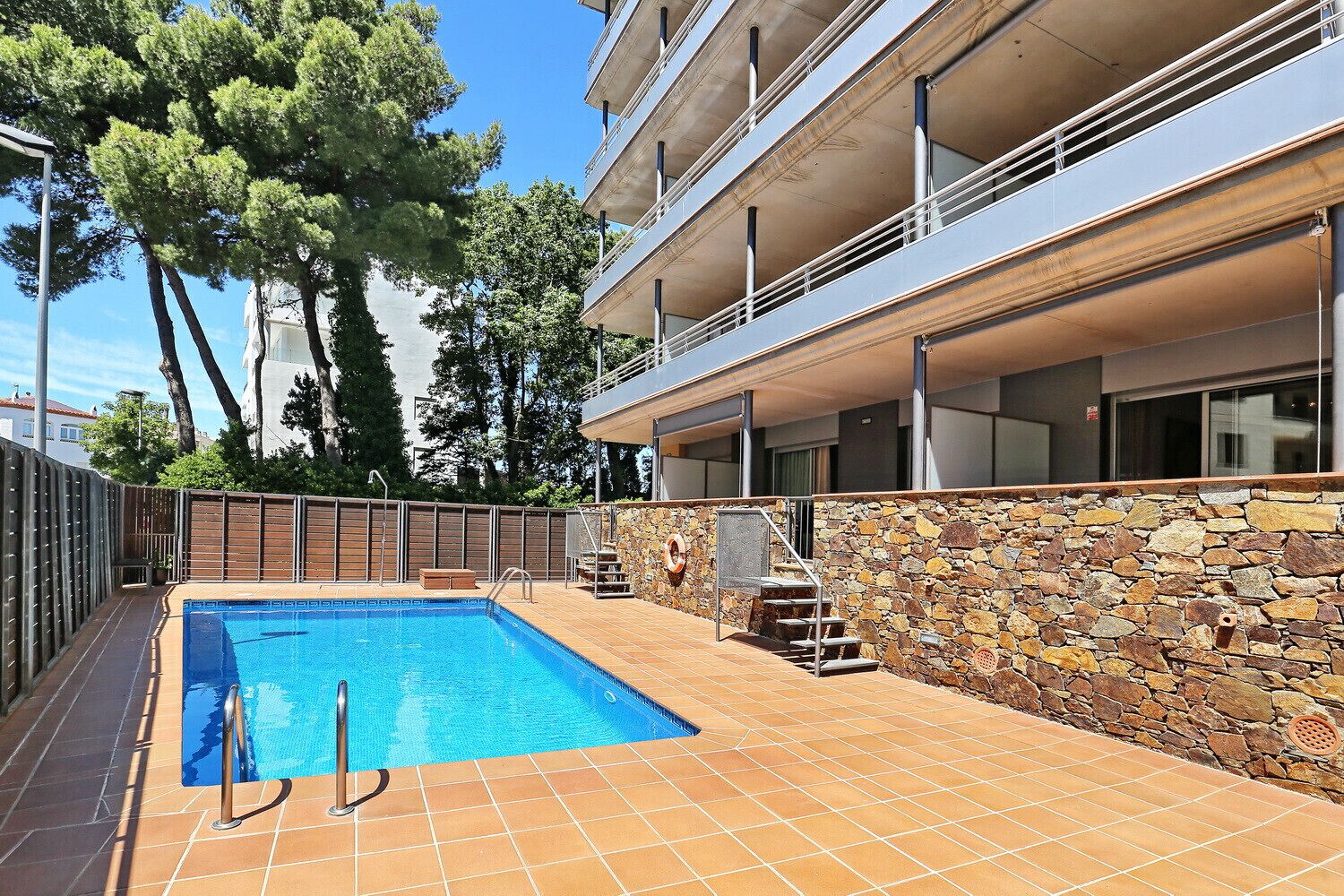 A swimming pool with a stone wall and a metal railing, surrounded by a fence and trees.