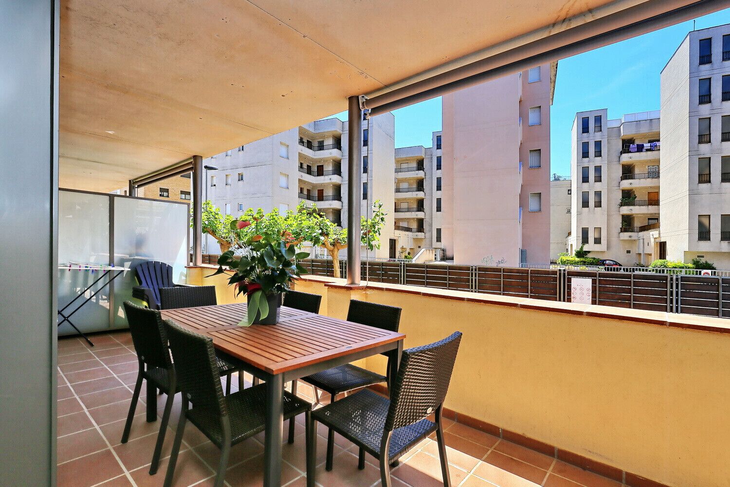 A balcony with a wooden table and chairs, a potted plant, and a view of apartment buildings.