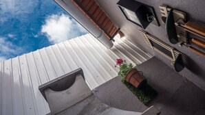 Una terraza en la azotea con techo de metal blanco, cubierta de madera, una planta en maceta con flores rosadas y mobiliario exterior.