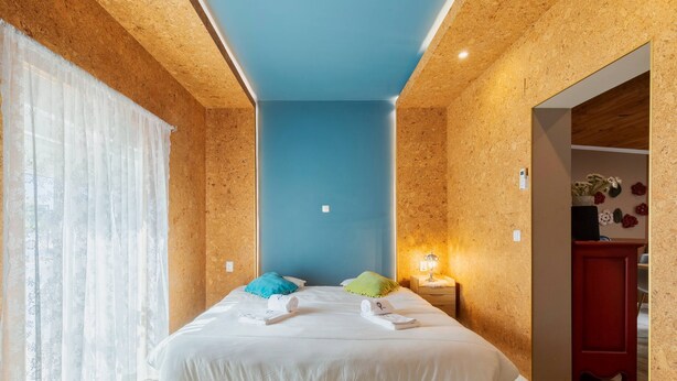 Un dormitorio con techo de madera, pared azul de acento, cama con ropa blanca y mesita de noche con lámpara.