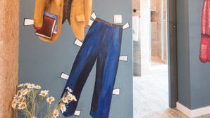 Una habitación con piso de madera, una pared blanca con aire acondicionado instalado, un estante blanco en la pared con una chaqueta amarilla y un libro, un jarrón con flores y una mesa de madera.