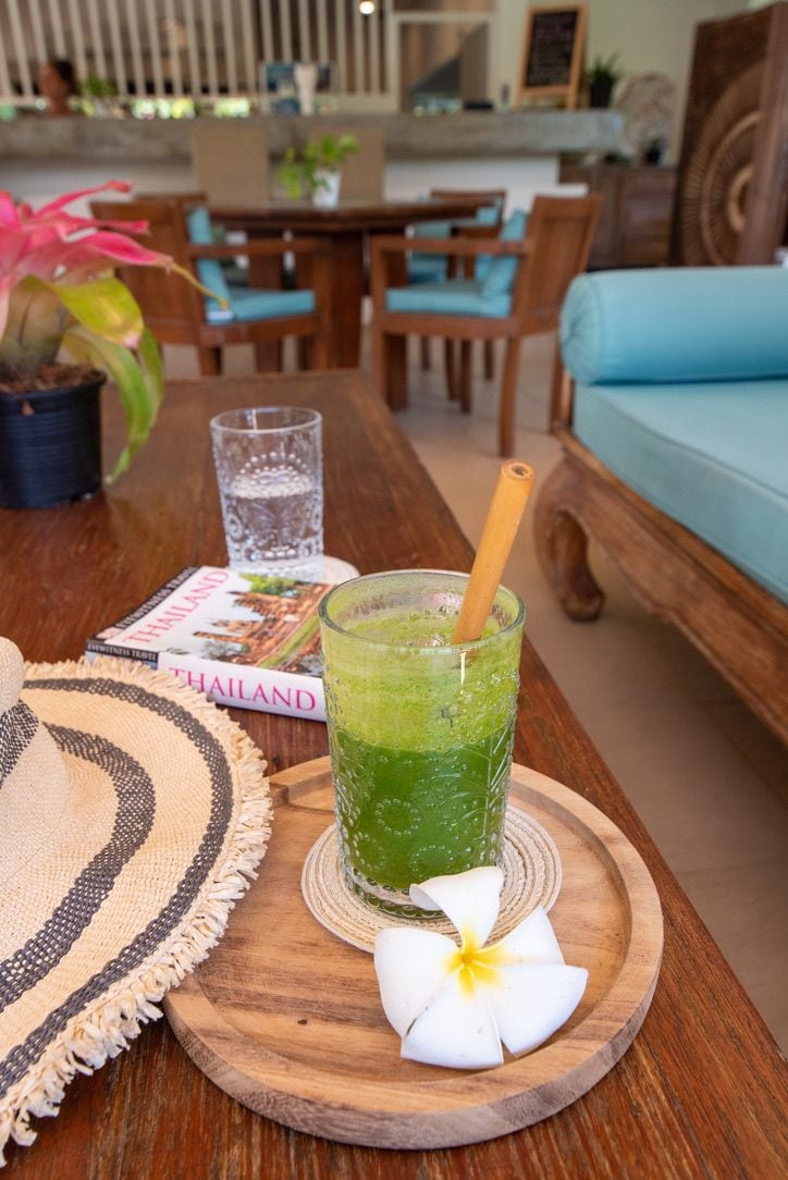 A wooden table with a glass of green drink, a book titled 'THAILAND,' and a woven placemat.