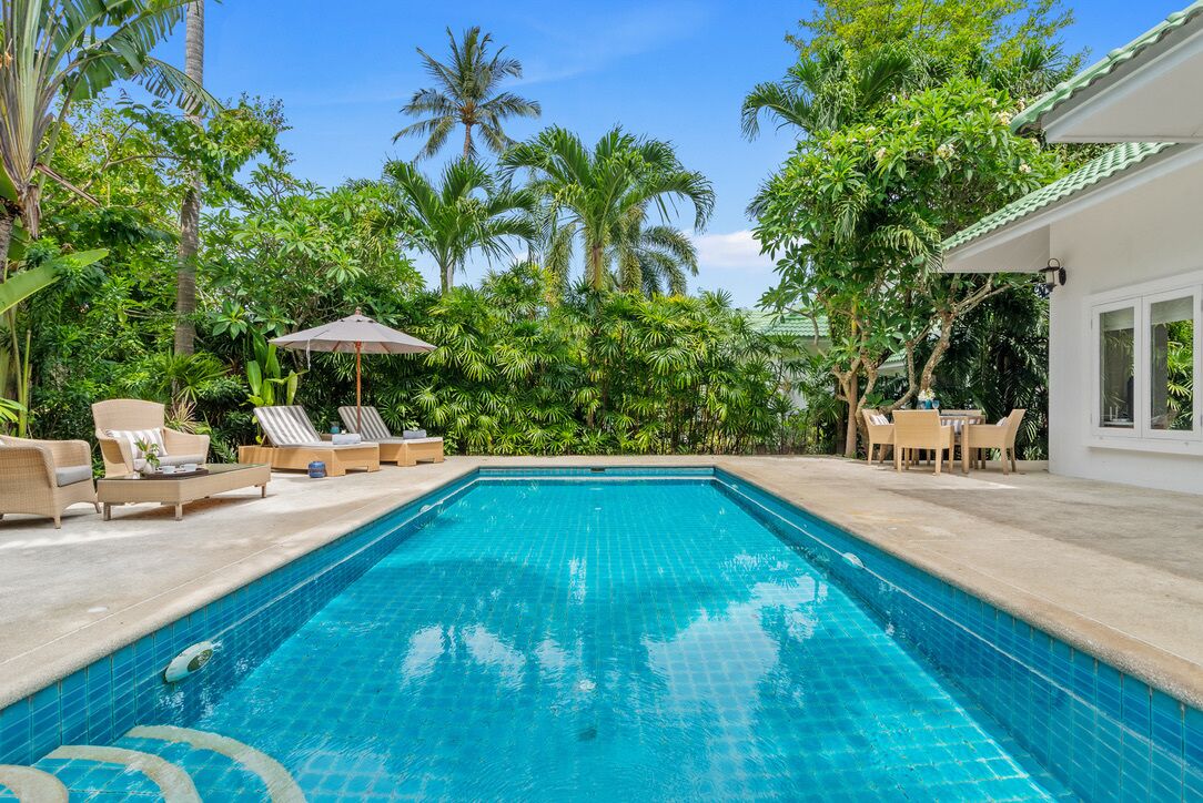 A rectangular swimming pool with clear blue water, surrounded by lounge chairs and umbrellas, with palm trees and a white building in the background.