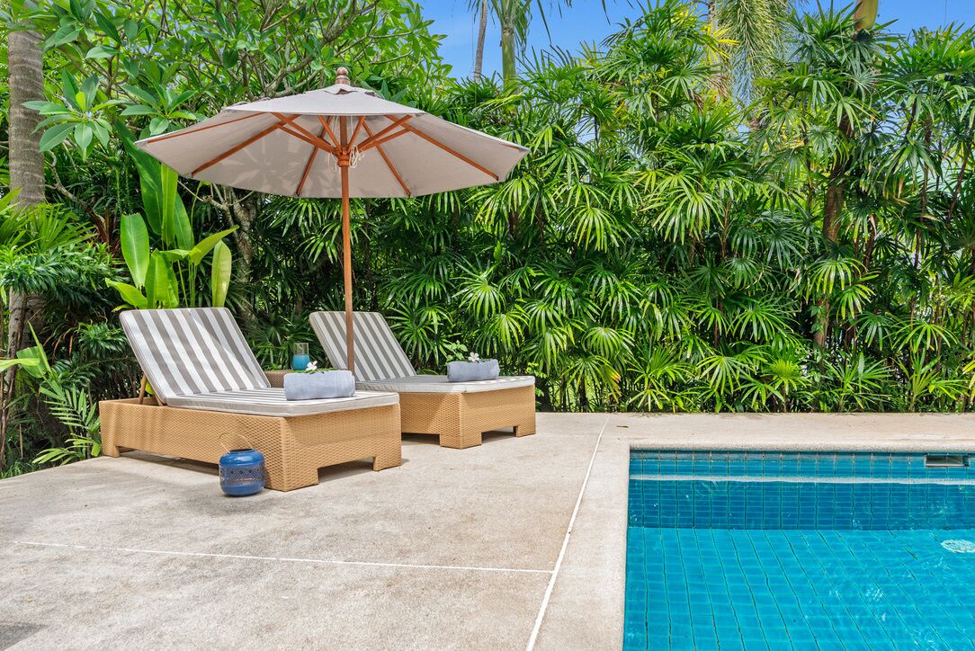 A poolside area with two lounge chairs and a large umbrella, surrounded by lush greenery.