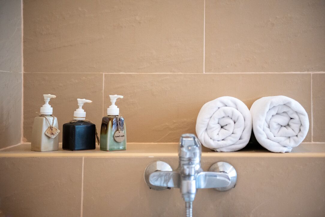 A bathroom with beige tiles, a shower with a chrome faucet, and three bottles of soap on a shelf.