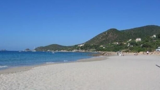 A beach with clear blue water, sandy shore, and a hill with buildings in the background.