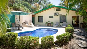 A pool area with lounge chairs and a table, surrounded by greenery and a building with a green roof. - Casa tropical - Located in Playa Guiones - Nosara - Costa Rica (Playa guiones)