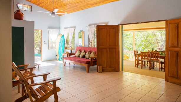 A spacious room with a wooden ceiling, a red sofa, wooden chairs, a dining table, and a view of greenery outside. - Casa tropical - Located in Playa Guiones - Nosara - Costa Rica (Playa guiones)