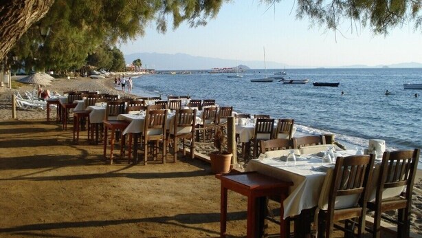 A seaside restaurant with outdoor seating, a view of the sea, and boats in the distance. - Private Pool , Wifi,BBQ, sea views Bodrum kadikalesi Garden Apartment (Bodrum Kadikalesi)
