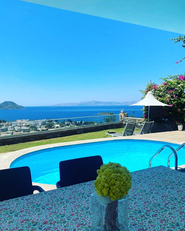 A pool with a view of the sea, a table with a floral tablecloth, and a vase with flowers.