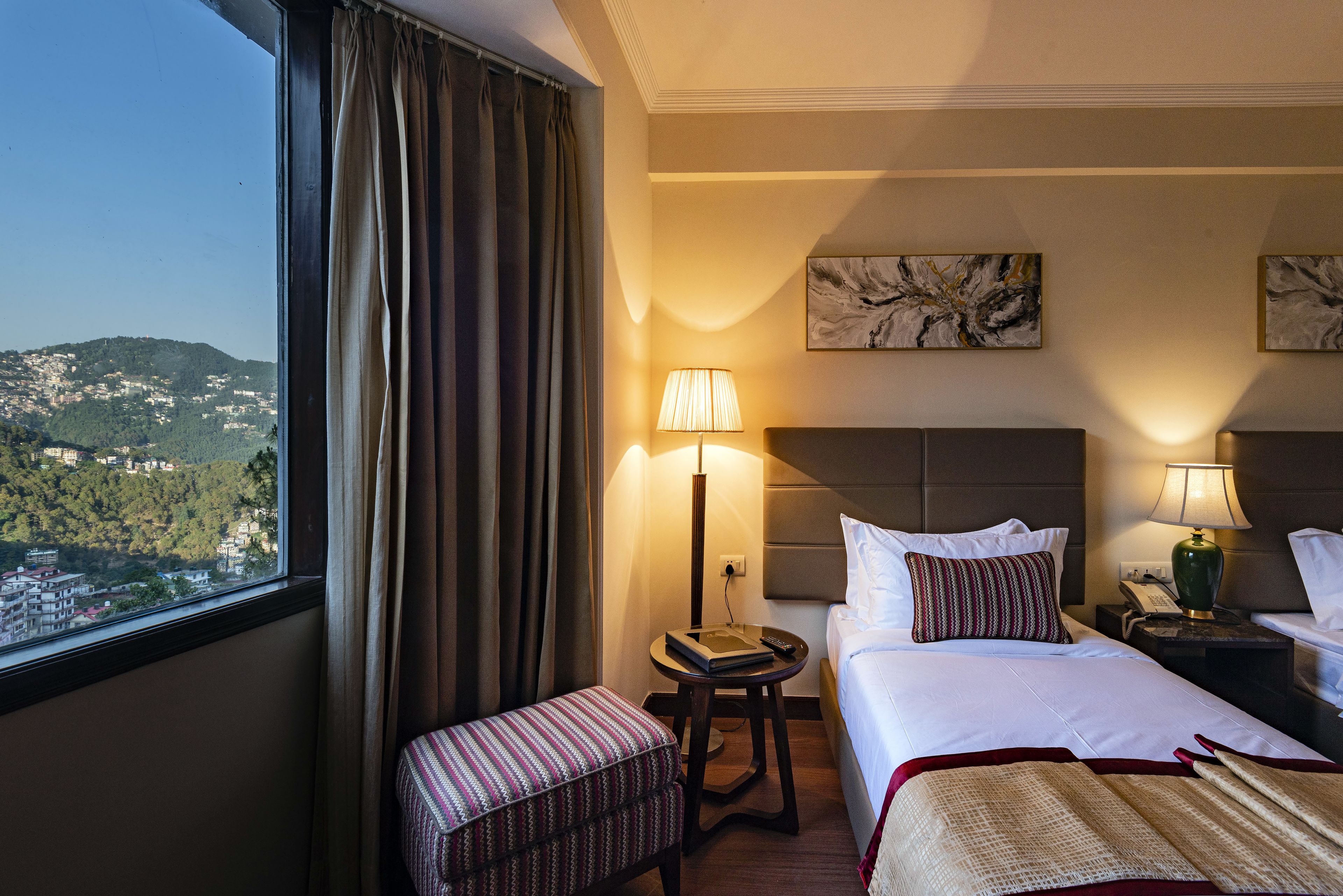 Een hotelkamer met een bed, nachtkastjes, een lamp, uitzicht op een bergachtig landschap door een raam, en een klein zithoekje.