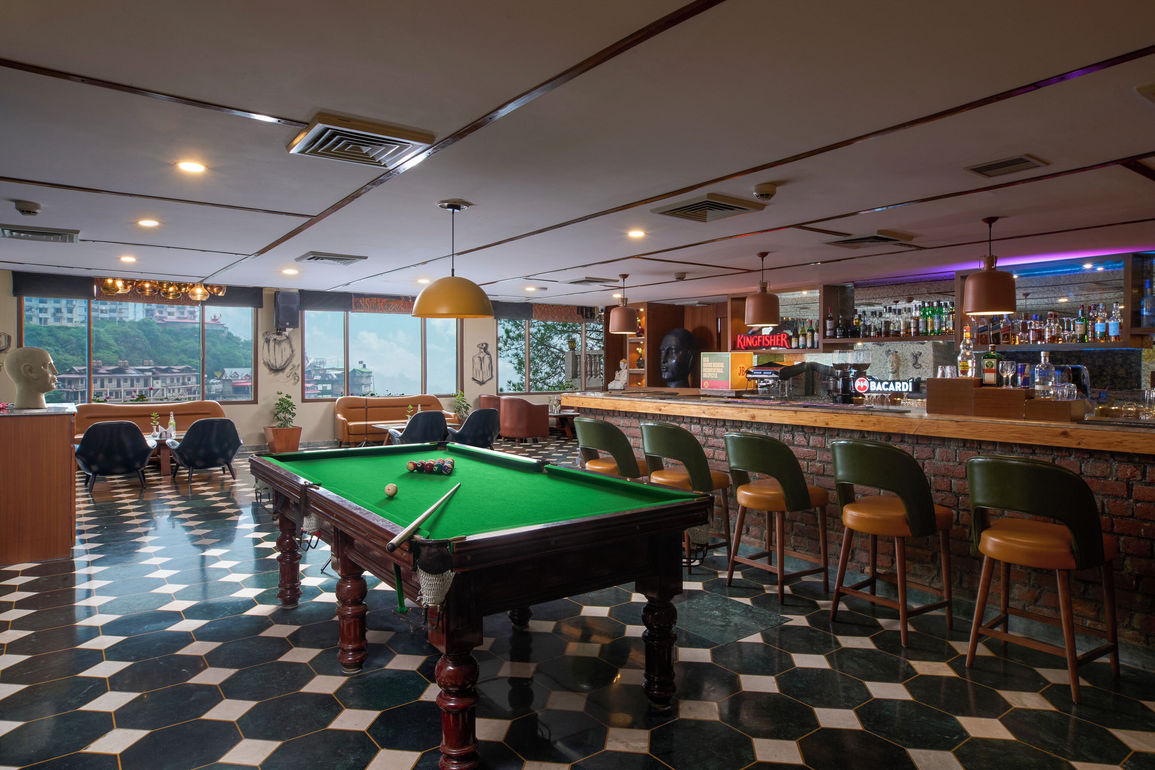 Una mesa de billar en una zona de bar con suelo a cuadros, taburetes y vistas a edificios a través de amplias ventanas.
