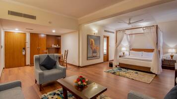 A hotel room with a bed, a sitting area with a chair and a table, a painting on the wall, and a ceiling fan.