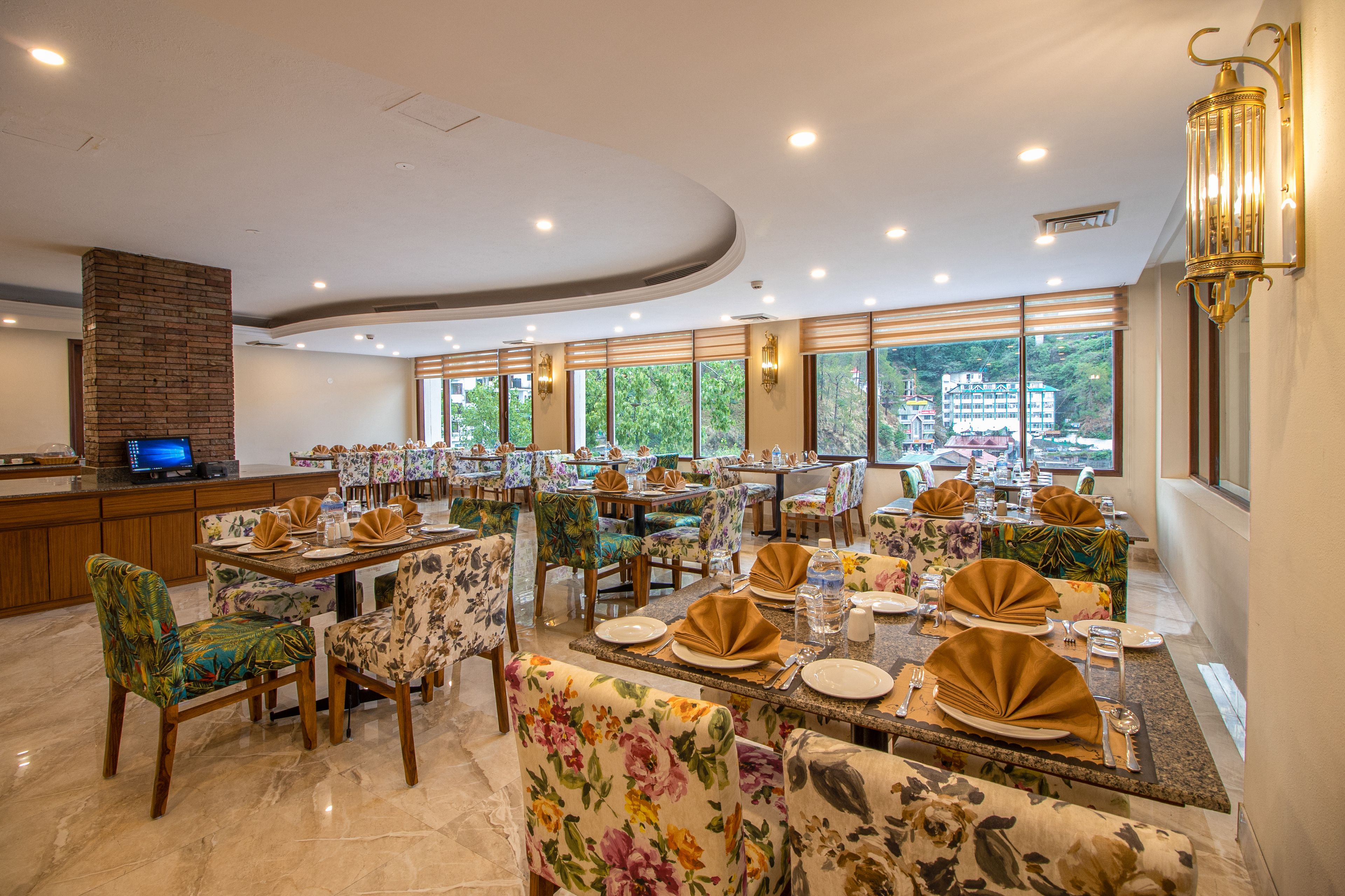 A restaurant with multiple dining tables set with gold napkins, floral chairs, and large windows offering a view of buildings and trees.