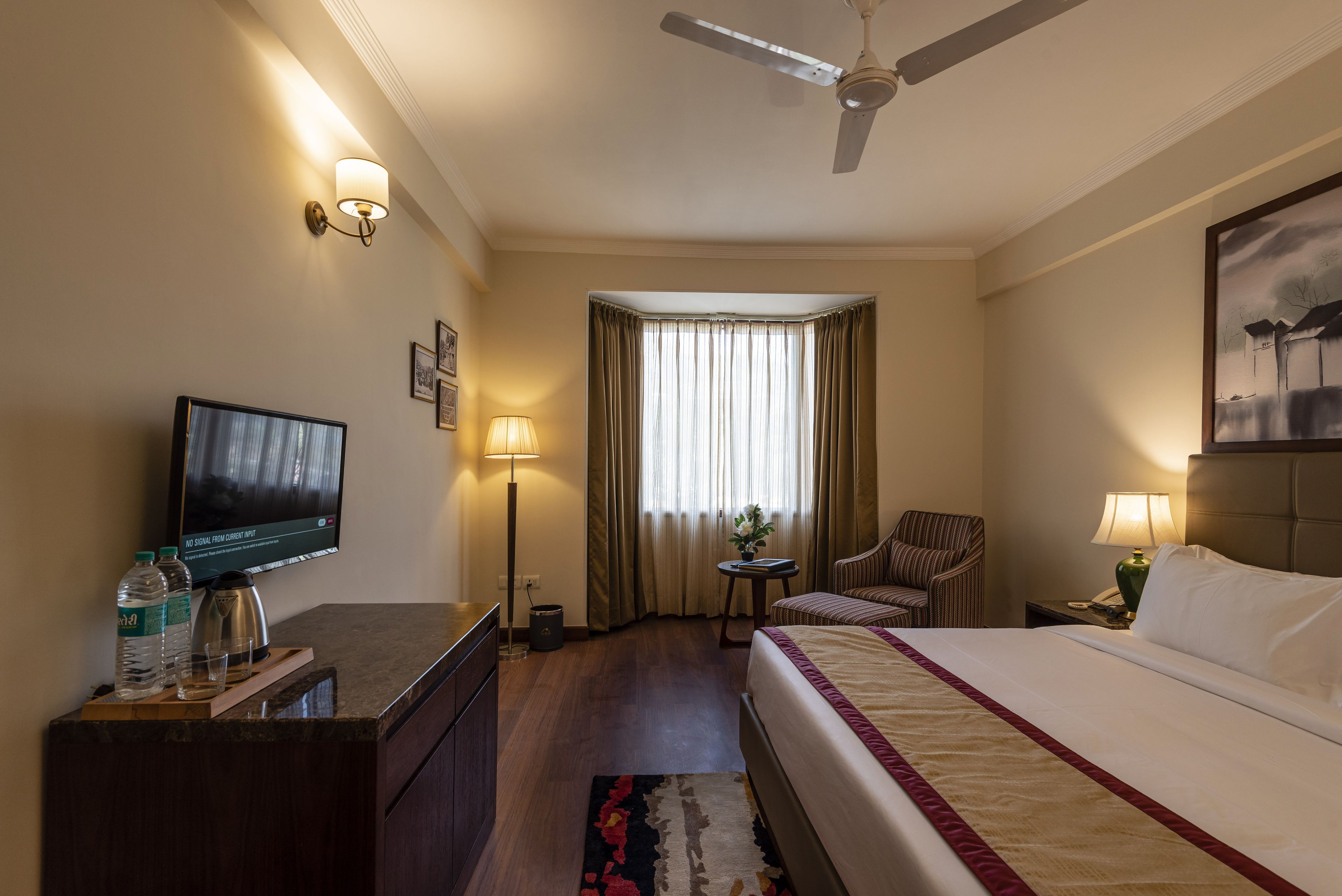 A hotel room with a bed, a television, a desk with a lamp, a chair, and a window with curtains.