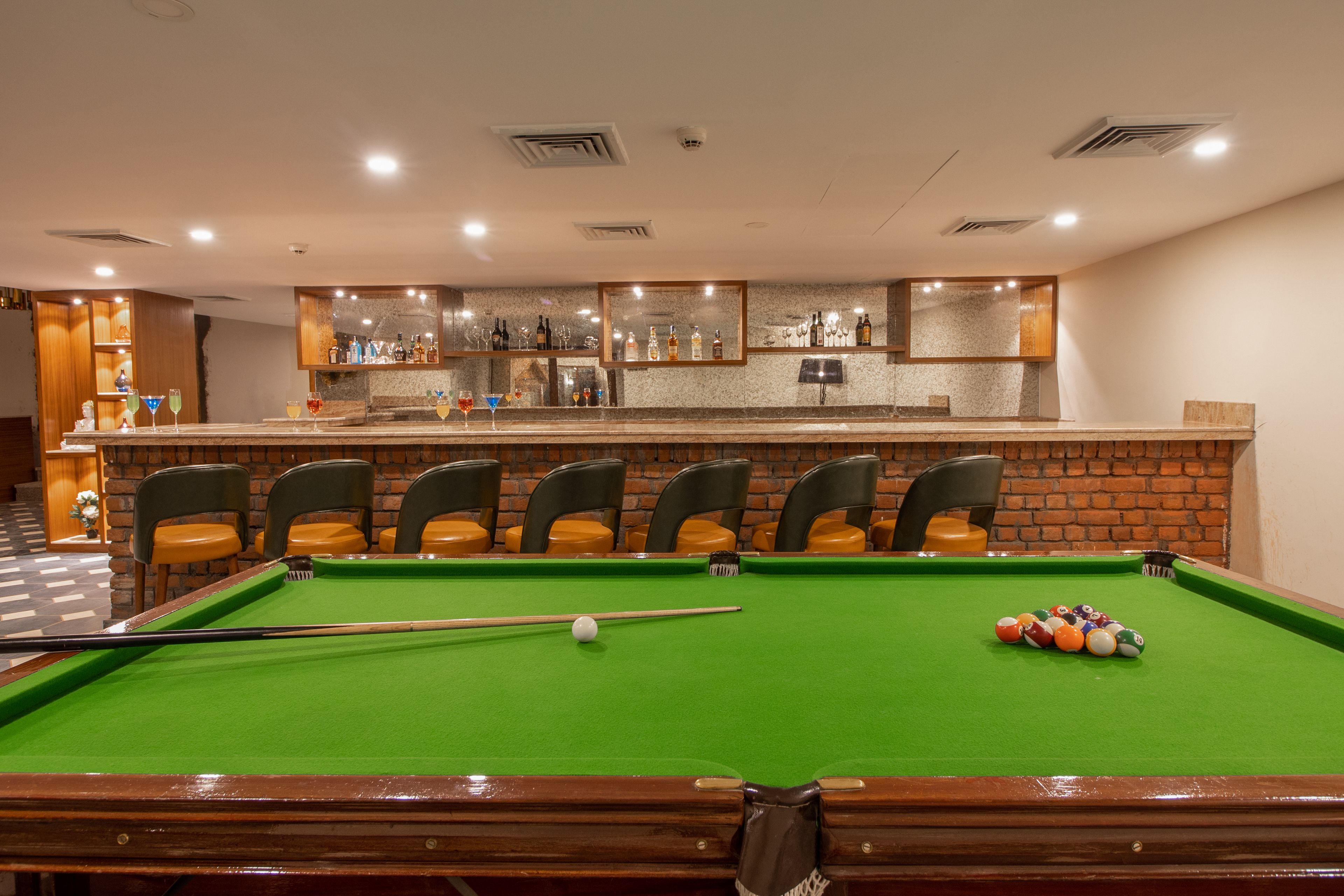 A pool table with a green felt surface, surrounded by four chairs. In the background, there is a bar area with a countertop displaying various bottles and glasses.