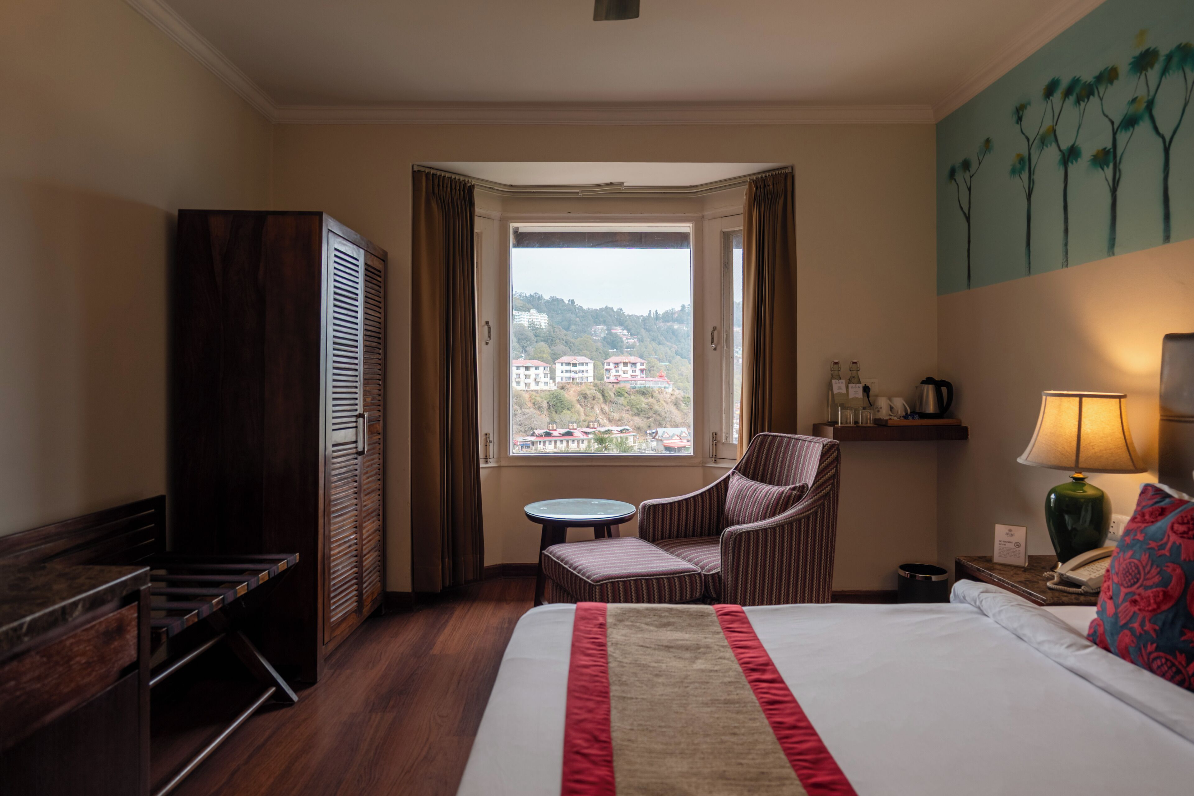 A hotel room with a large bed, a chair, a small table, a wardrobe, a window with a view of buildings, and a painting on the wall.