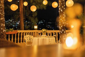 Un área de comedor al aire libre con una mesa preparada para dos personas, rodeada de árboles y luces colgantes.