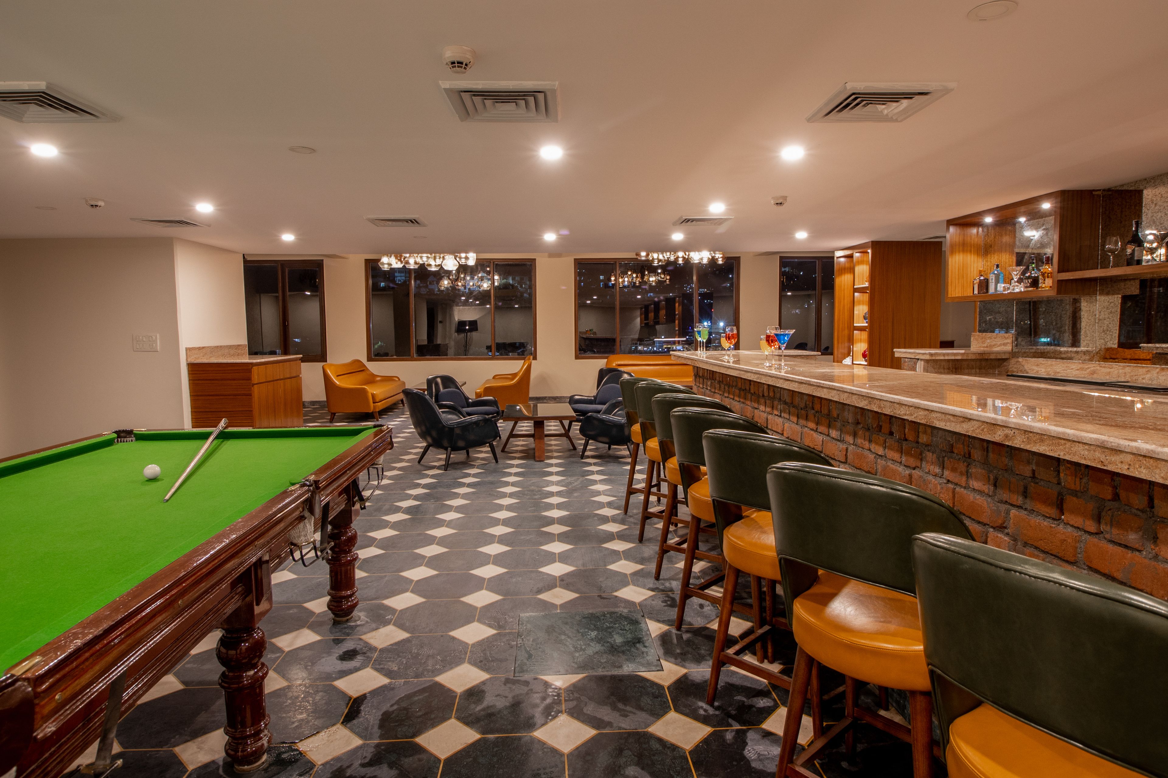Una mesa de billar con taco y bolas, rodeada de taburetes de bar y una zona de bar con mostrador y estanterías.