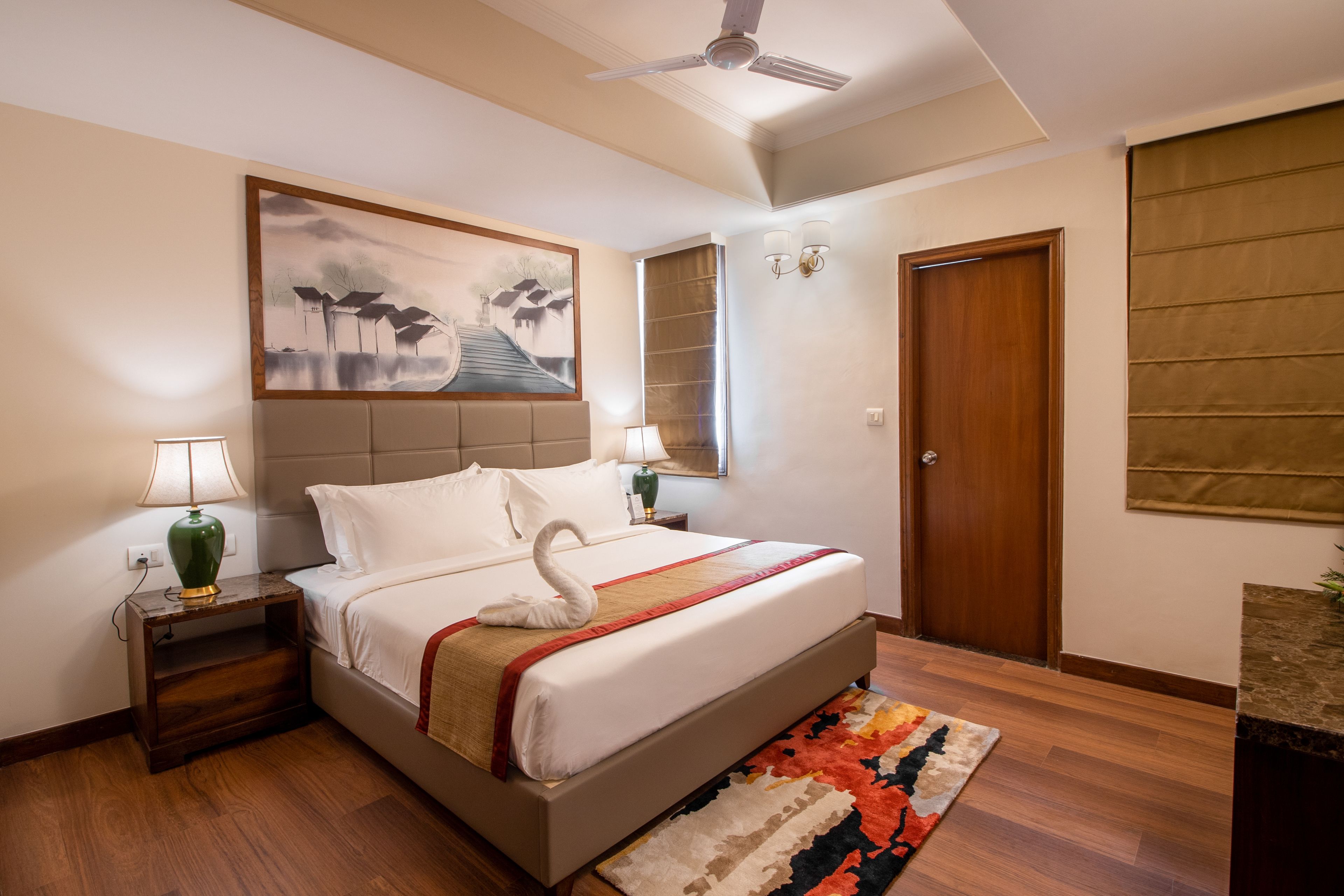 A hotel room with a bed, a nightstand with a lamp, a wall-mounted painting, a door, and a ceiling fan.