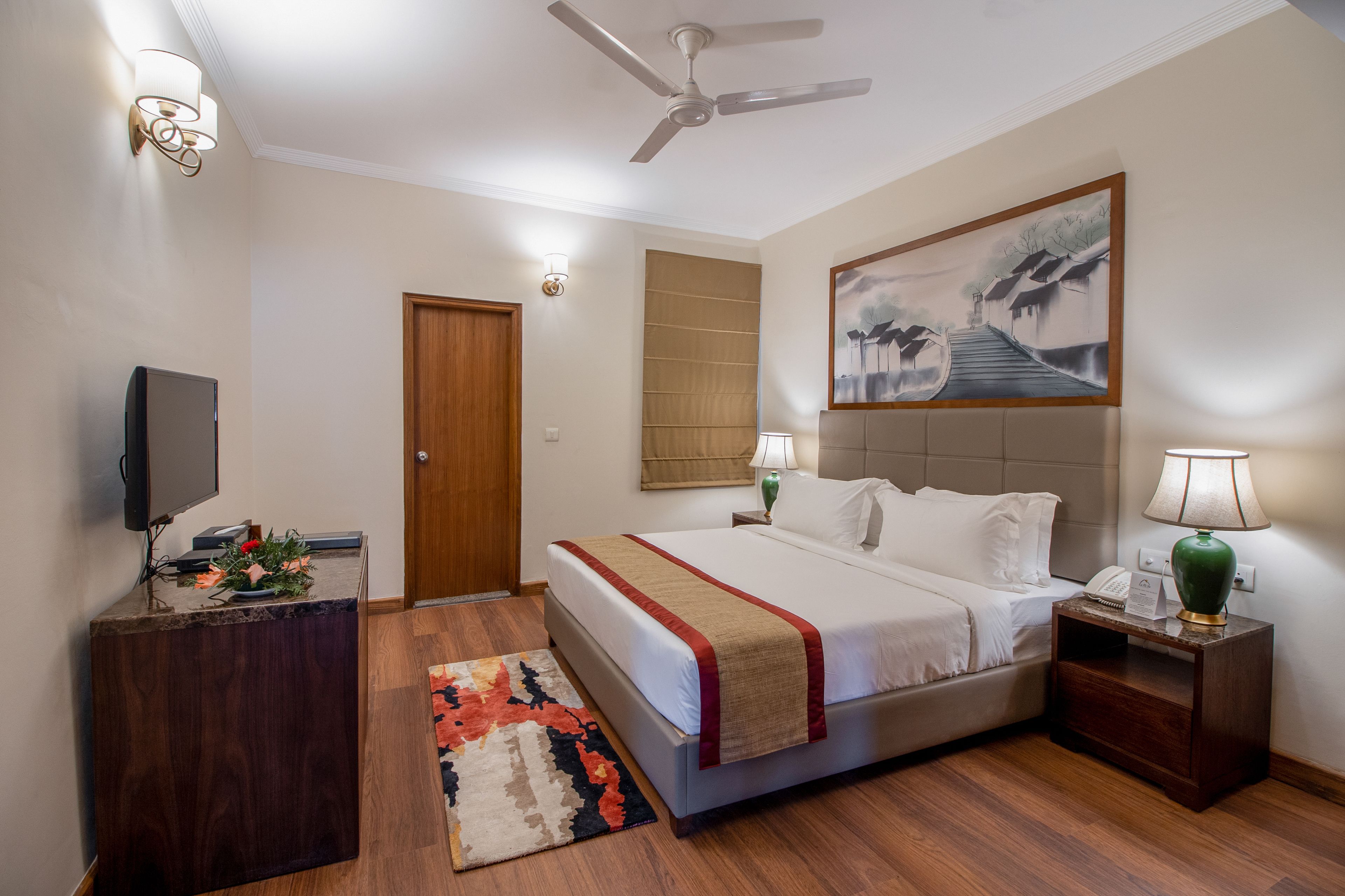 Quarto de hotel com cama, mesas de cabeceira, escrivaninha, televisão, ventoinha no teto e um quadro na parede.