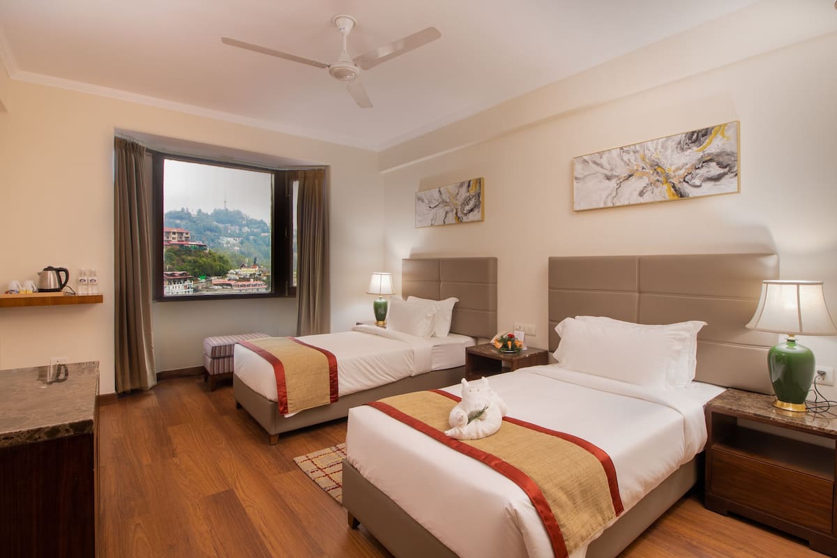 Une chambre d’hôtel avec deux lits, un bureau, une chaise, une fenêtre avec des rideaux, un ventilateur de plafond et des œuvres d’art accrochées aux murs.