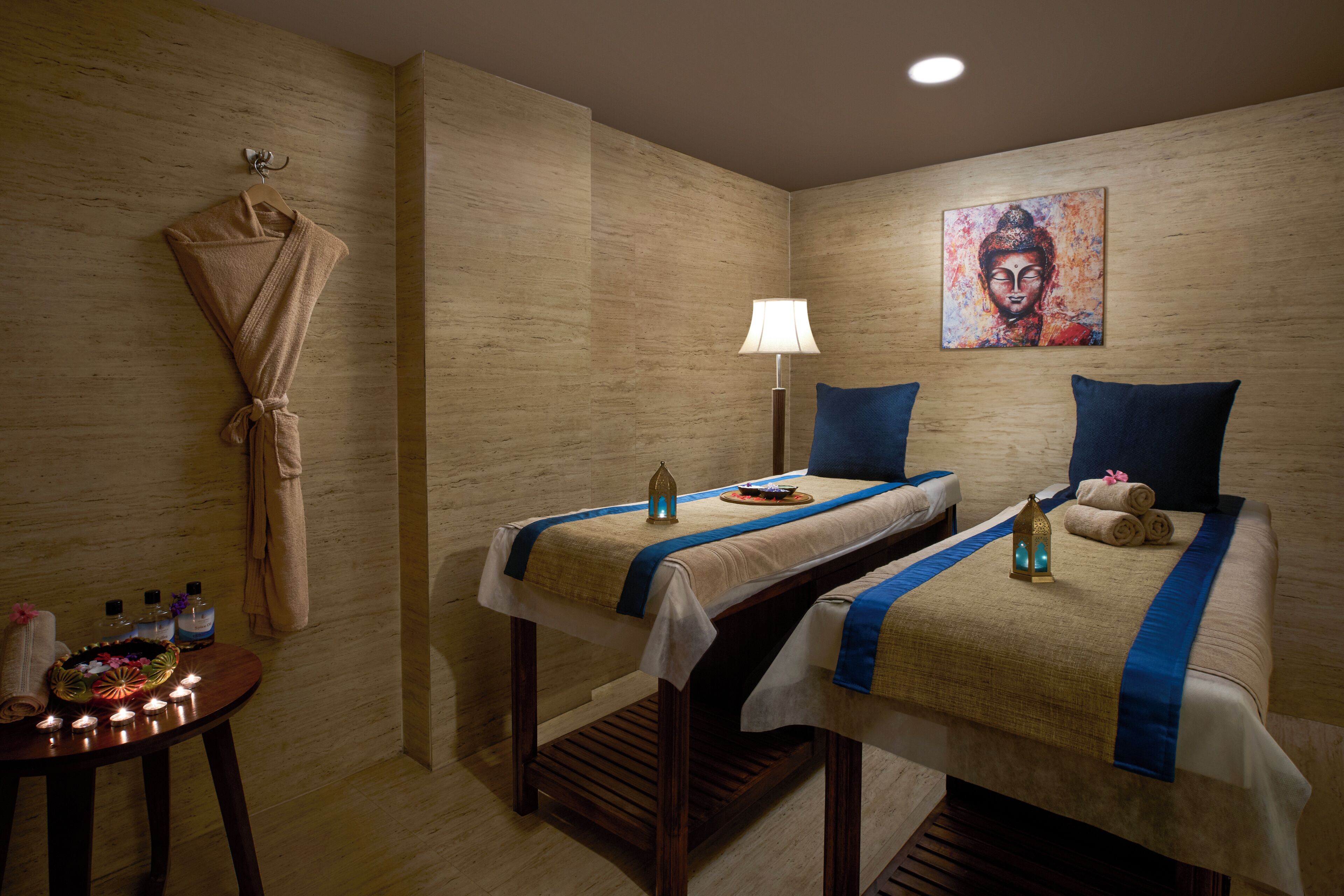 A room with two beds, a wall-mounted robe rack, a small table with candles and toiletries, and a painting on the wall.