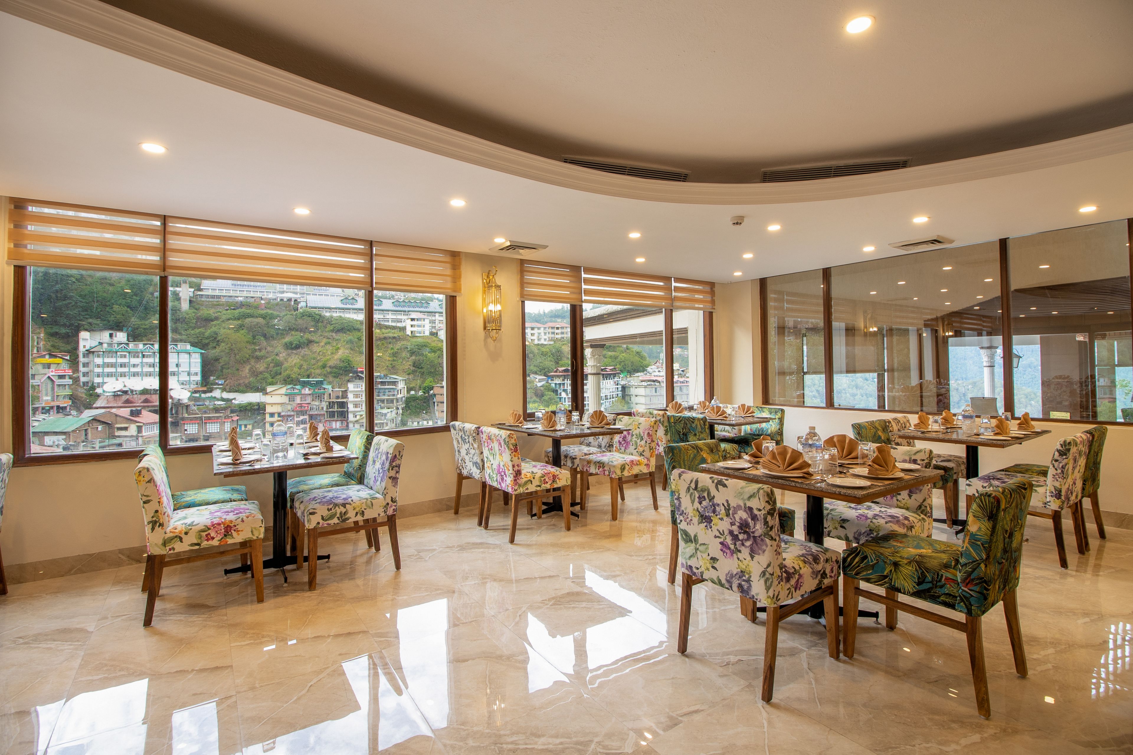A restaurant with multiple tables and chairs, large windows with city views, and a polished floor.