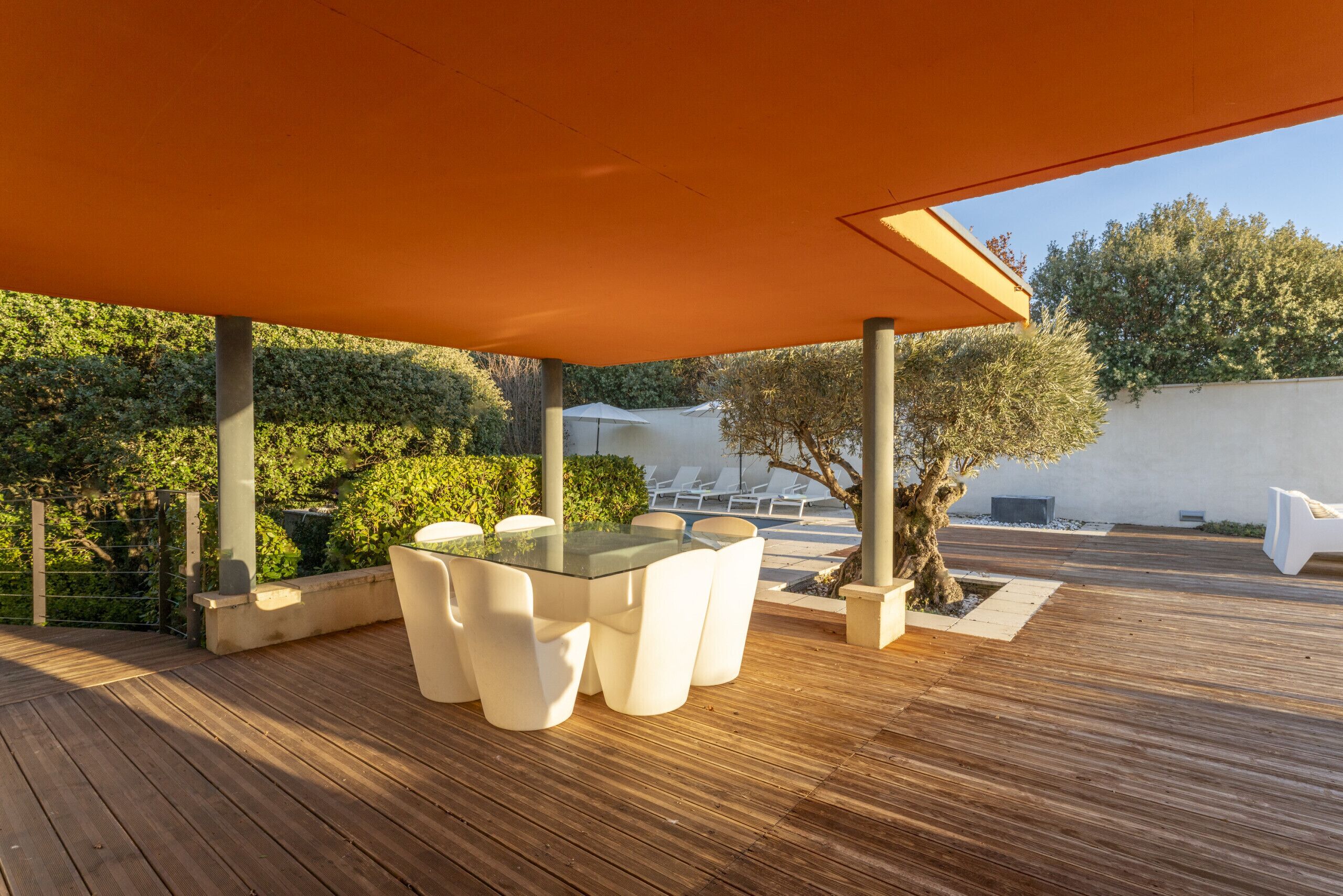 An outdoor patio with a glass-top table and white chairs, surrounded by wooden decking and greenery.