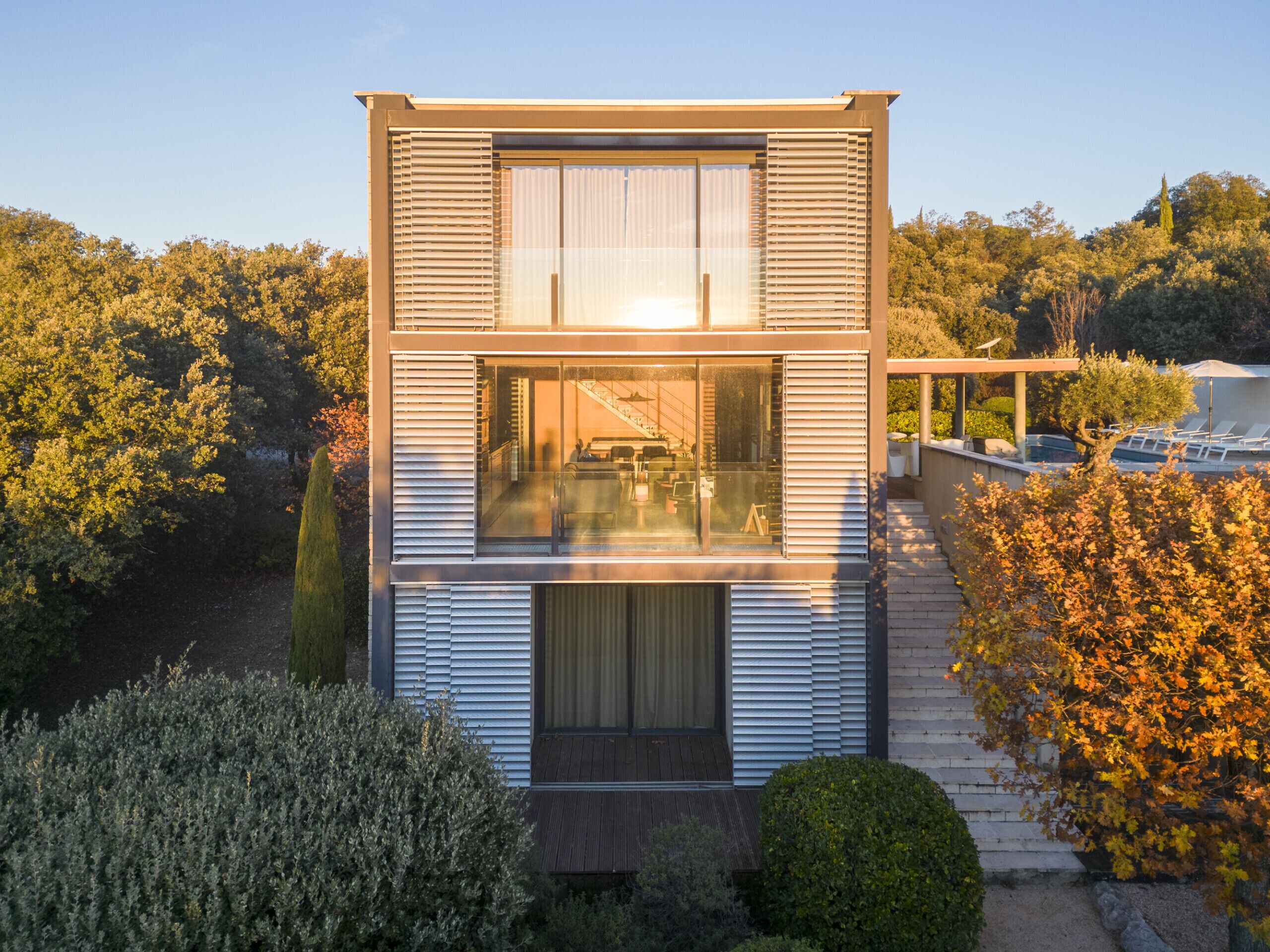 A modern two-story house with large windows, a covered outdoor area, and a view of the surrounding trees.