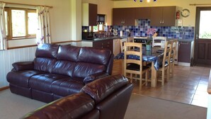 A living room with a leather sofa, a dining area with wooden chairs, a kitchen with blue tiles, and a wooden ceiling.