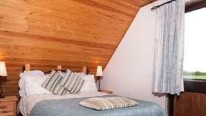 A bedroom with a sloped ceiling, a bed with striped bedding, two bedside lamps, a wooden nightstand, and a view of the outdoors through a window with curtains.