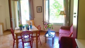Una habitación con una mesa de comedor, sillas, un sofá, una mesa de centro con un jarrón de flores y una ventana con cortinas.