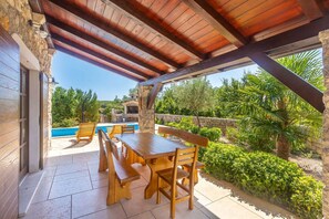 Une terrasse extérieure avec une table et des bancs en bois, un mur en pierre et une vue sur une piscine et la verdure.