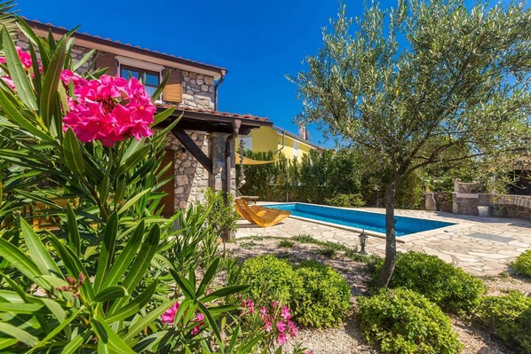 Un espace aménagé au bord d’une piscine en pierre, avec un hamac et un bâtiment en pierre dont la façade est jaune.
