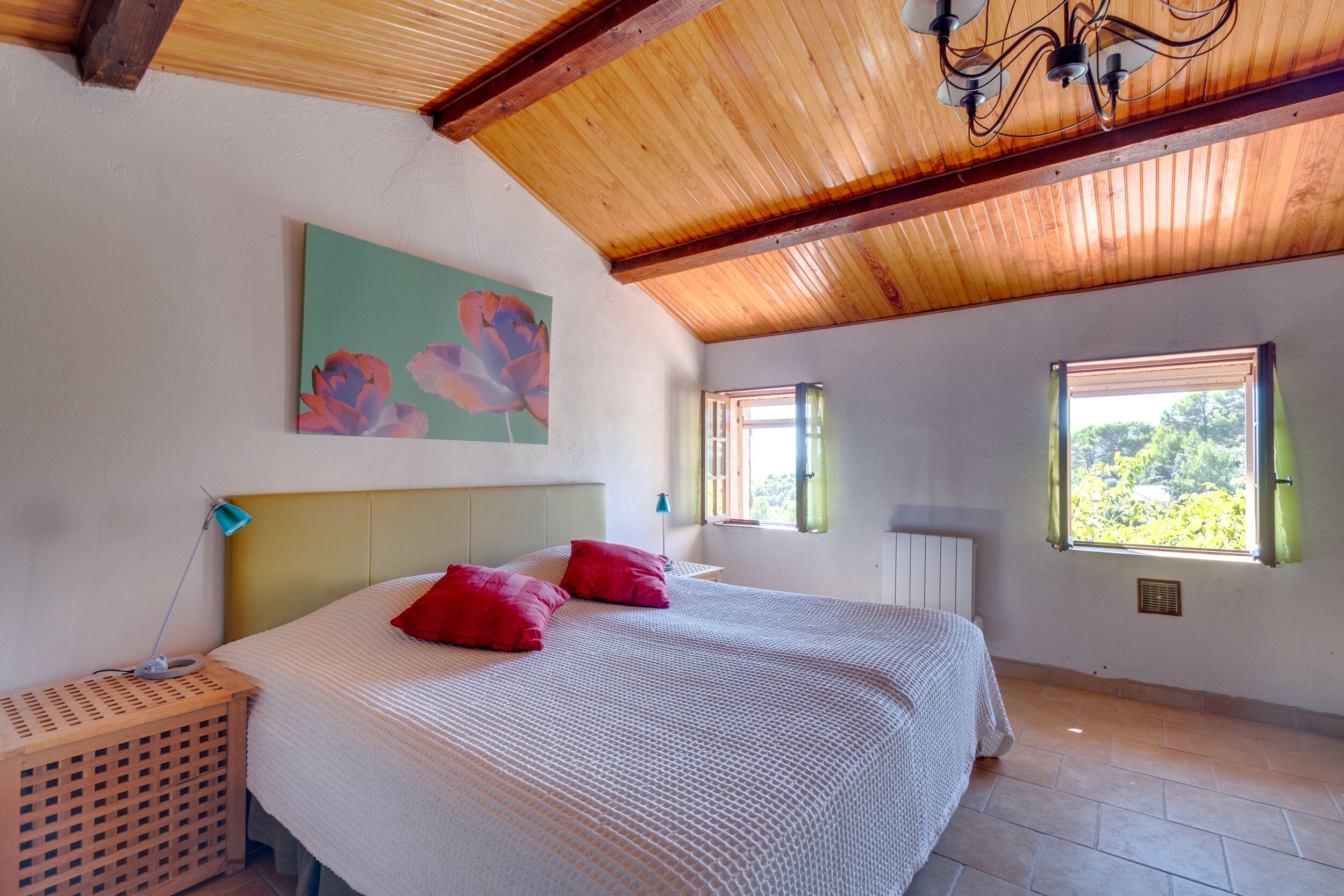 Een slaapkamer met een houten plafond, een bed met witte beddengoed en rode kussens, een schilderij van bloemen, en een raam met uitzicht op groen.