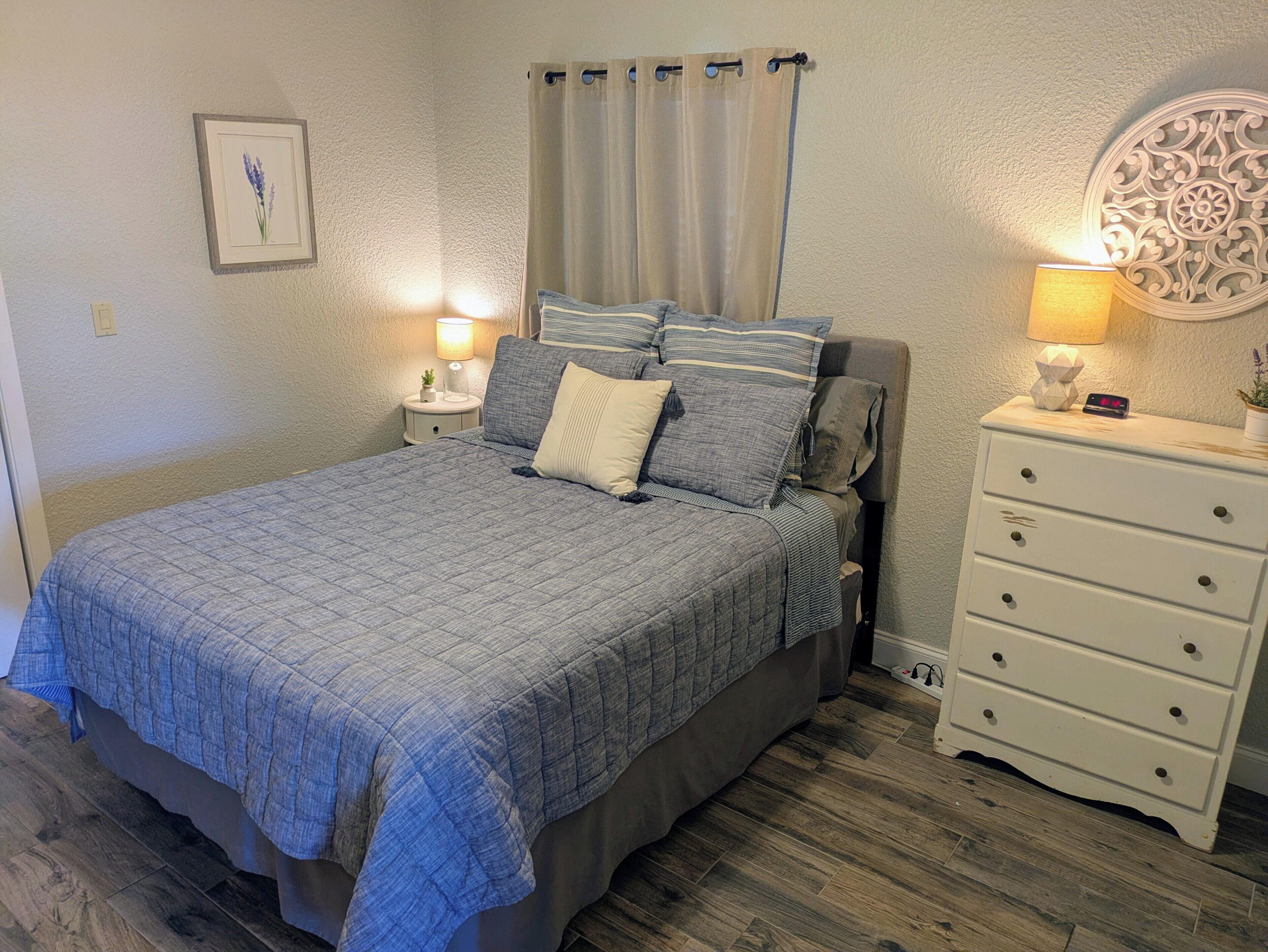 A bedroom with a bed, nightstands, a dresser, a wall-mounted lamp, a framed picture, and a curtain.