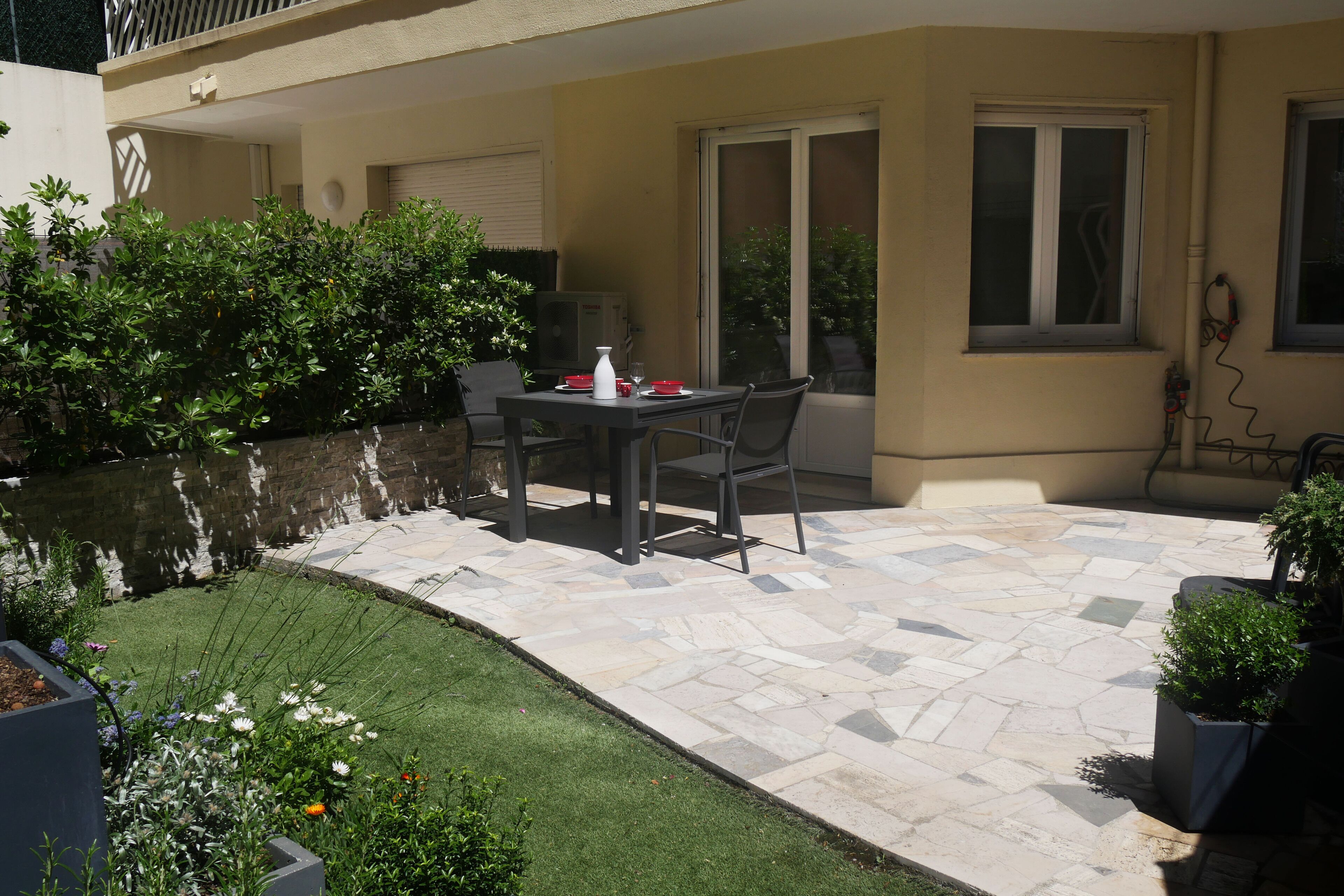 Une terrasse extérieure avec un sol en pierre, une table entourée de chaises et un petit jardin agrémenté d’un bassin.