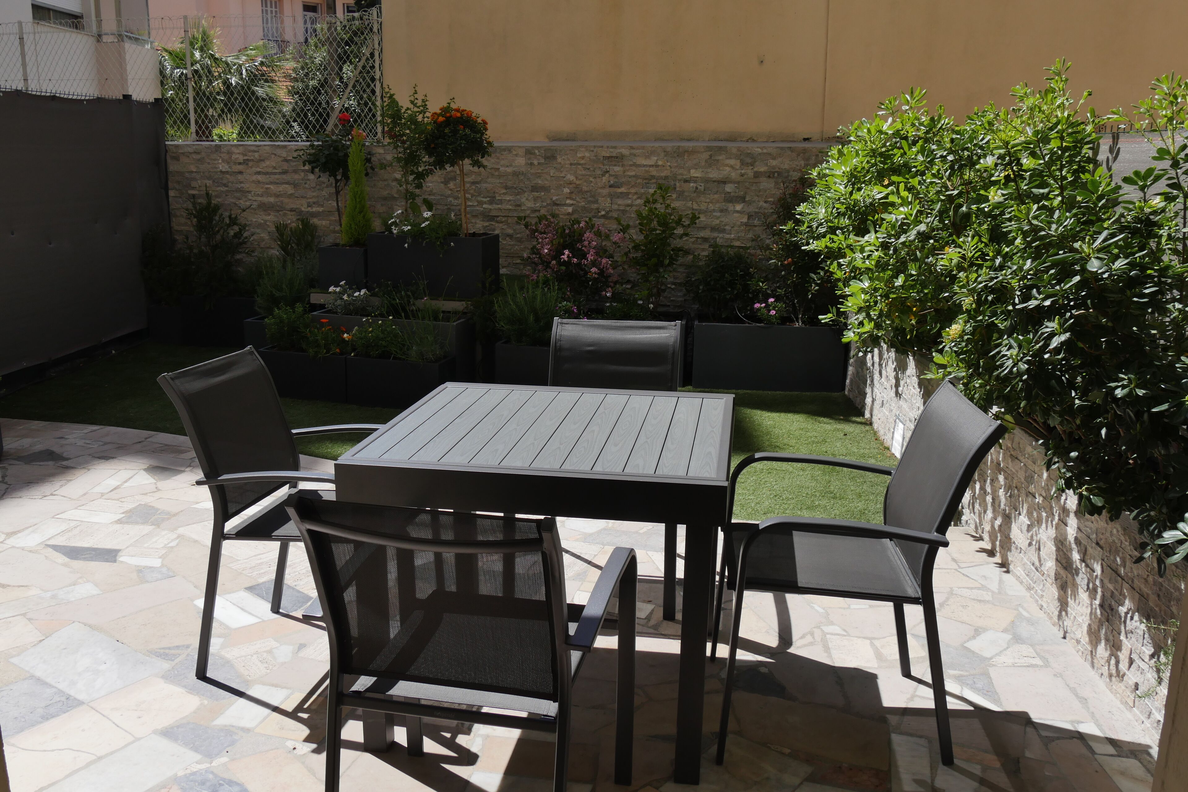 Un patio exterior con una mesa y sillas, rodeado de plantas en macetas y un muro de piedra.