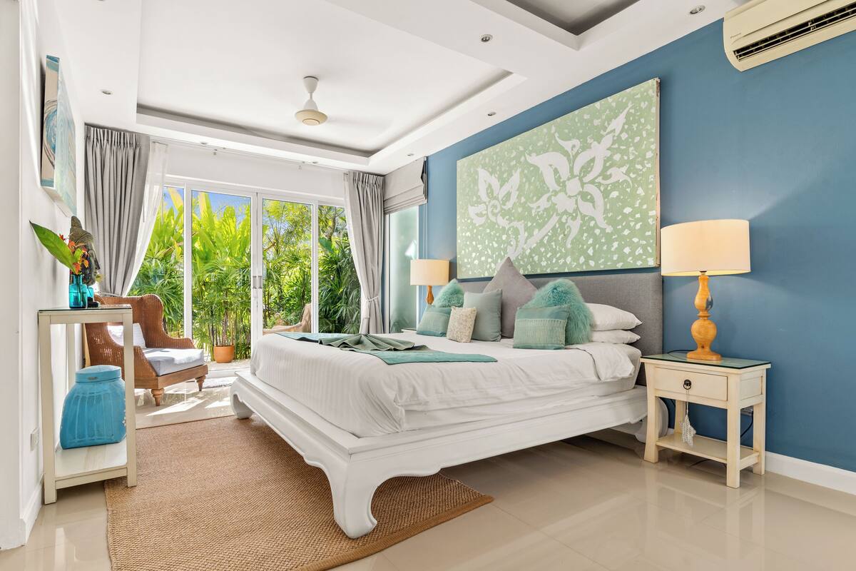 A bedroom with a large bed, a nightstand, a chair, and a view of greenery outside.