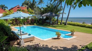 A rectangular swimming pool with a clear blue water, surrounded by a concrete deck. There are lounge chairs and a blue umbrella providing shade. The pool area is adjacent to a well-maintained lawn and palm trees. - Baan Rim Haad (Koh Samui)